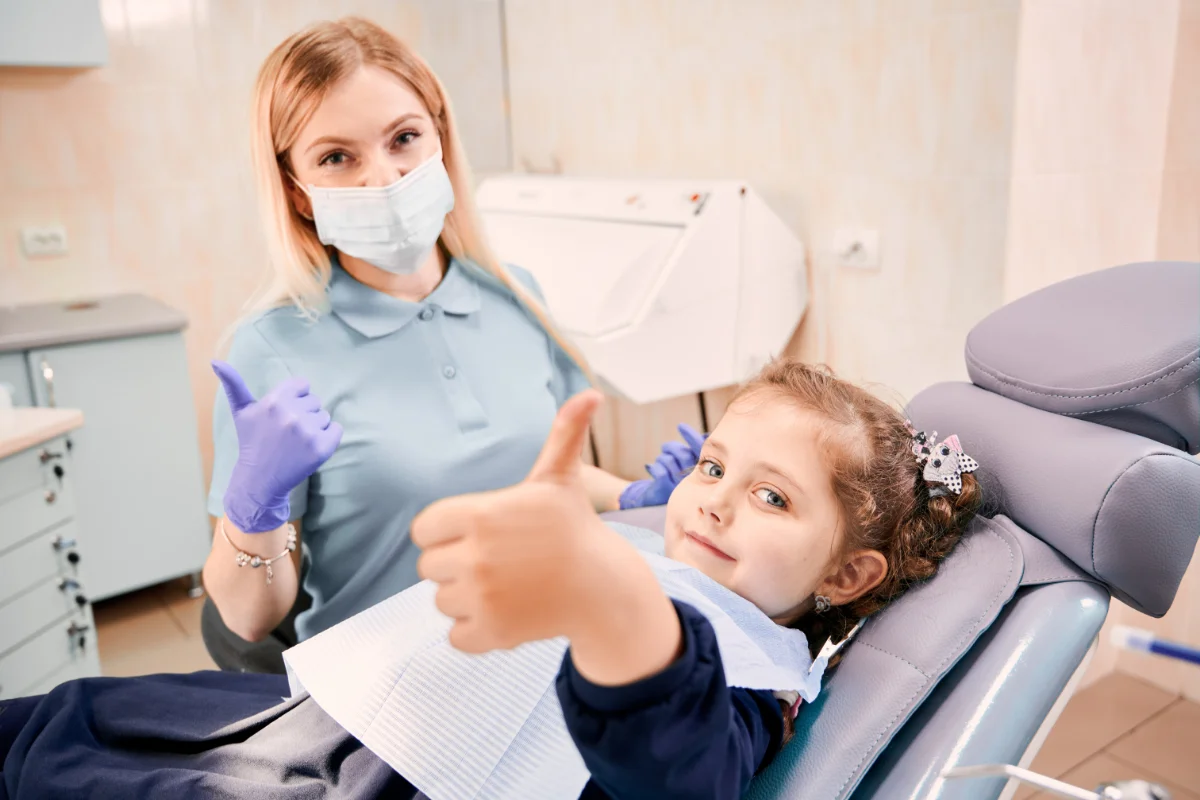 A happy little girl giving a thumbs up while sitting in a dental chair next to a female pediatric dentist in Vienna, VA wearing gloves and a face mask